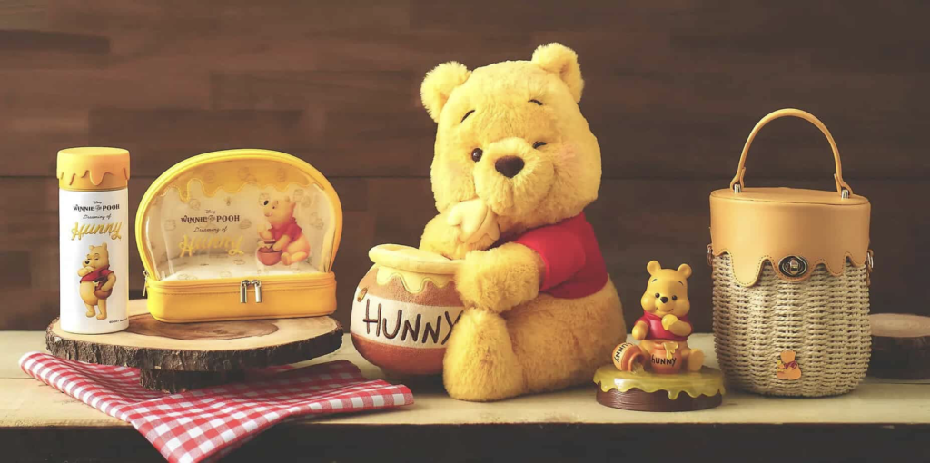 Une peluche Winnie l'Ourson est assise avec un pot "Hunny", entourée d'articles à thème comprenant un gobelet, une pochette, une figurine et un panier sur une surface en bois avec un tissu à carreaux rouges - parfait pour les fans depuis la sortie du film le 4 Février 1966.