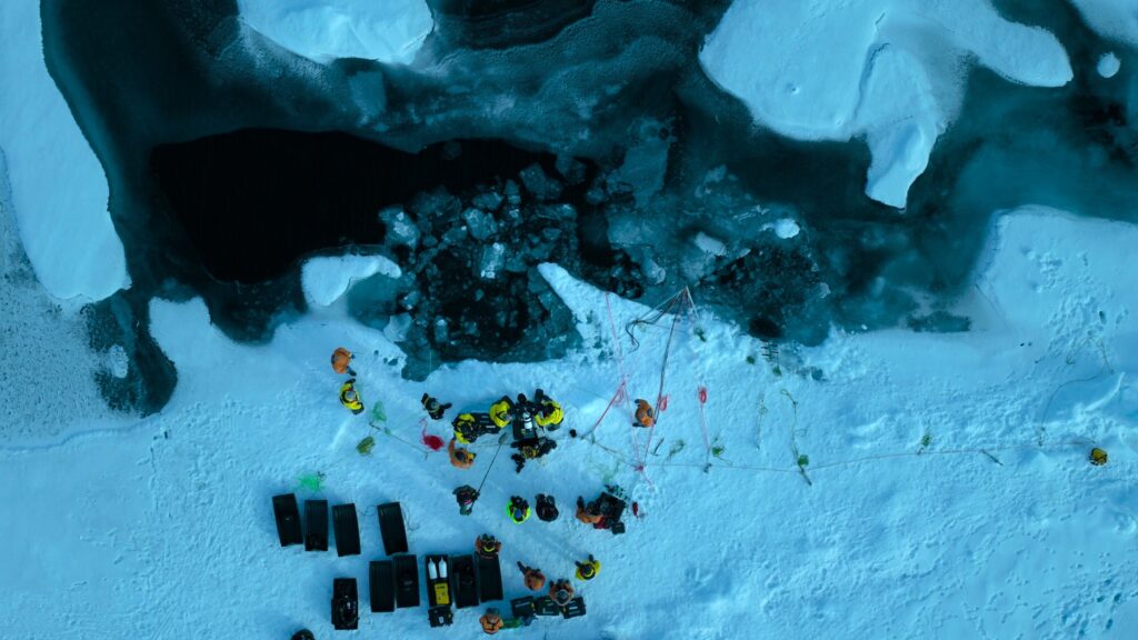 Vue aérienne de personnes en gilet jaune et orange rassemblées autour d'un équipement sur un sol enneigé près d'une étendue d'eau partiellement gelée, rappelant les explorations de Nat Geo présentées sur Disney+.