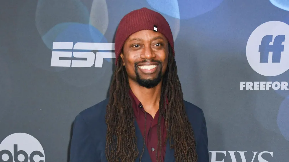 Un homme avec de longues dreadlocks et un bonnet marron sourit devant une toile de fond avec les logos d'ESPN et de Freeform, rappelant Saladin Patterson lors d'un événement organisé par la 20e chaîne de télévision pour King Of The Hill.