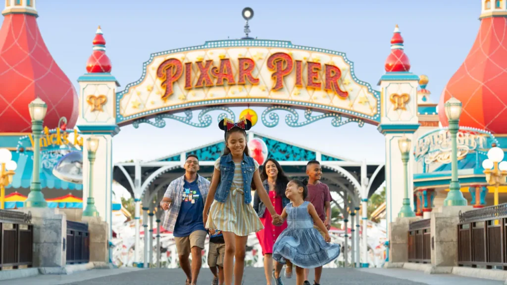Une famille se promène sous le panneau d'entrée de Pixar Pier à Disney California Adventure, avec des décorations et des attractions colorées en arrière-plan, mettant en valeur la magie créée par les créateurs de Disney.