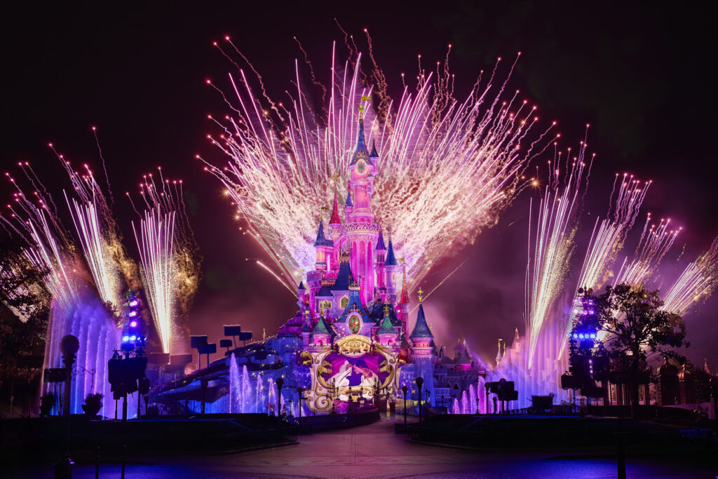 Un château illuminé de nuit avec des feux d'artifice colorés explosant dans le ciel derrière lui, entouré de fontaines illuminées dans l'un des parcs emblématiques de Disney.