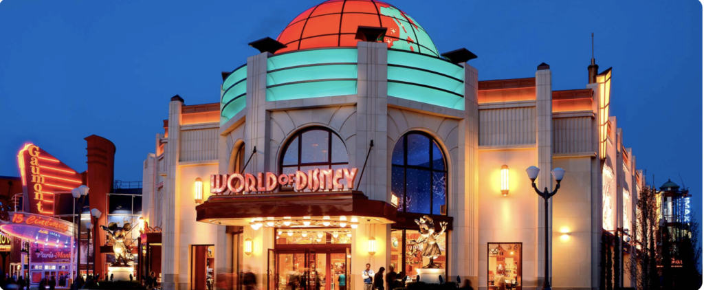 Shopping. Vue extérieure d'une boutique World of Disney éclairée de nuit, avec un dôme illuminé, des fenêtres cintrées et des enseignes colorées. Idéal pour acheter des souvenirs de Disneyland Paris après une journée magique au parc Disneyland Paris.