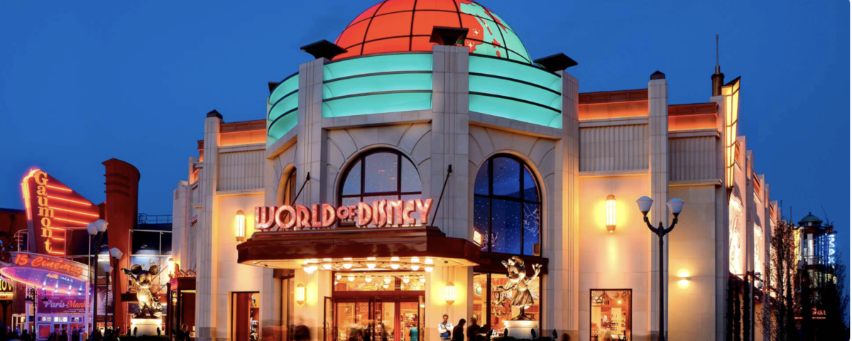 Vue extérieure d'une boutique World of Disney éclairée de nuit, avec un dôme illuminé, des fenêtres cintrées et des enseignes colorées. Idéal pour acheter des souvenirs de Disneyland Paris après une journée magique au parc Disneyland Paris.
