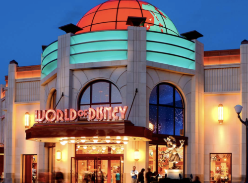 Vue extérieure d'une boutique World of Disney éclairée de nuit, avec un dôme illuminé, des fenêtres cintrées et des enseignes colorées. Idéal pour acheter des souvenirs de Disneyland Paris après une journée magique au parc Disneyland Paris.