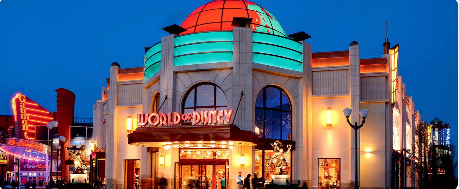 Vue extérieure d'une boutique World of Disney éclairée de nuit, avec un dôme illuminé, des fenêtres cintrées et des enseignes colorées. Idéal pour acheter des souvenirs de Disneyland Paris après une journée magique au parc Disneyland Paris.
