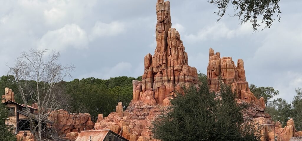 Une formation rocheuse rougeâtre ressemblant à une montagne s'élève derrière des bâtiments en bois rustiques et des arbres sous un ciel nuageux, évoquant la frontière sauvage du chemin de fer Big Thunder Mountain Railroad du Magic Kingdom.