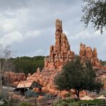 Une formation rocheuse rougeâtre ressemblant à une montagne s'élève derrière des bâtiments en bois rustiques et des arbres sous un ciel nuageux, évoquant la frontière sauvage du chemin de fer Big Thunder Mountain Railroad du Magic Kingdom.