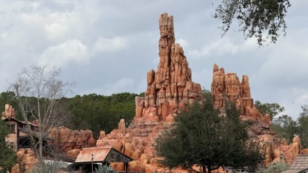 Une formation rocheuse rougeâtre ressemblant à une montagne s'élève derrière des bâtiments en bois rustiques et des arbres sous un ciel nuageux, évoquant la frontière sauvage du chemin de fer Big Thunder Mountain Railroad du Magic Kingdom.