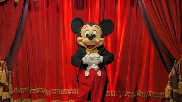 Une personne déguisée en Mickey Mouse se tient devant des rideaux de scène rouges fermés et ornés de franges dorées à Disneyland Paris, parfaits pour des photos mémorables et des conseils pour éviter les files d'attente.