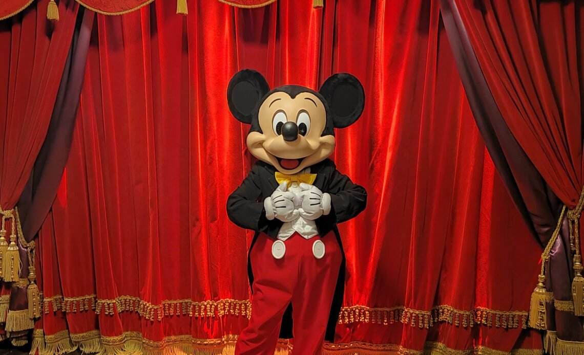 Une personne déguisée en Mickey Mouse se tient devant des rideaux de scène rouges fermés et ornés de franges dorées à Disneyland Paris, parfaits pour des photos mémorables et des conseils pour éviter les files d'attente.