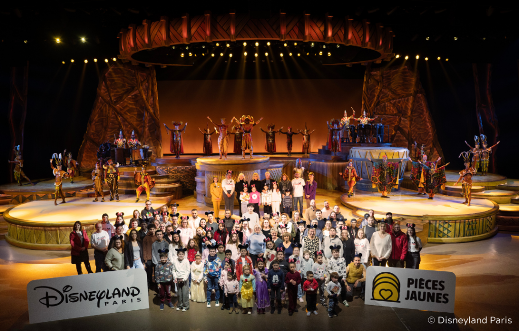 Un grand groupe de personnes pose sur la scène de Disneyland Paris avec des artistes en costume ; les logos de Disneyland Paris et de Pièces Jaunes 2026 sont visibles au premier plan, célébrant le lancement des produits solidaires.