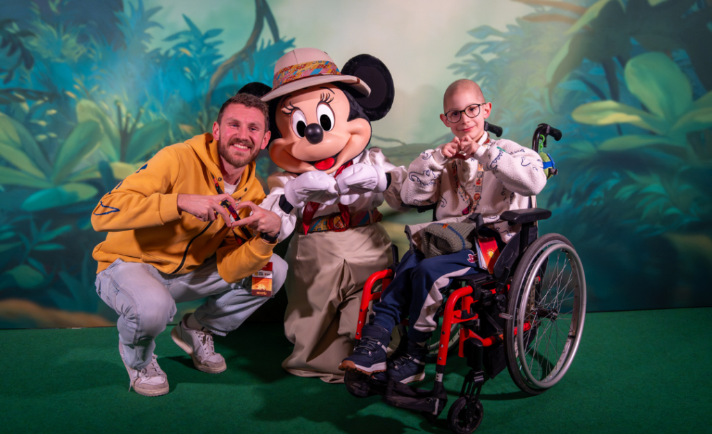 Un homme, une personne en fauteuil roulant et un personnage costumé posent ensemble pour une photo dans un décor de jungle à Disneyland Paris lors de l'événement Pièces Jaunes 2026 pour les enfants hospitalisés.