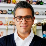 Le directeur marketing de Disney, Asad Ayaz, portant des lunettes, se tient devant des étagères présentant des figurines de collection et plusieurs récompenses en or.