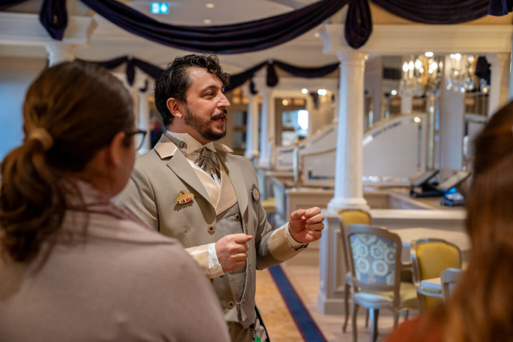 Un homme en costume beige s'adresse à deux personnes dans une salle décorée et élégante avec des colonnes, des chaises et des tissus drapés lors d'un événement de recrutement à Villeurbanne.