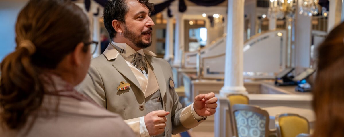 Un homme en costume beige s'adresse à deux personnes dans une salle décorée et élégante avec des colonnes, des chaises et des tissus drapés lors d'un événement de recrutement à Villeurbanne.