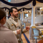 Un homme en costume beige s'adresse à deux personnes dans une salle décorée et élégante avec des colonnes, des chaises et des tissus drapés lors d'un événement de recrutement à Villeurbanne.