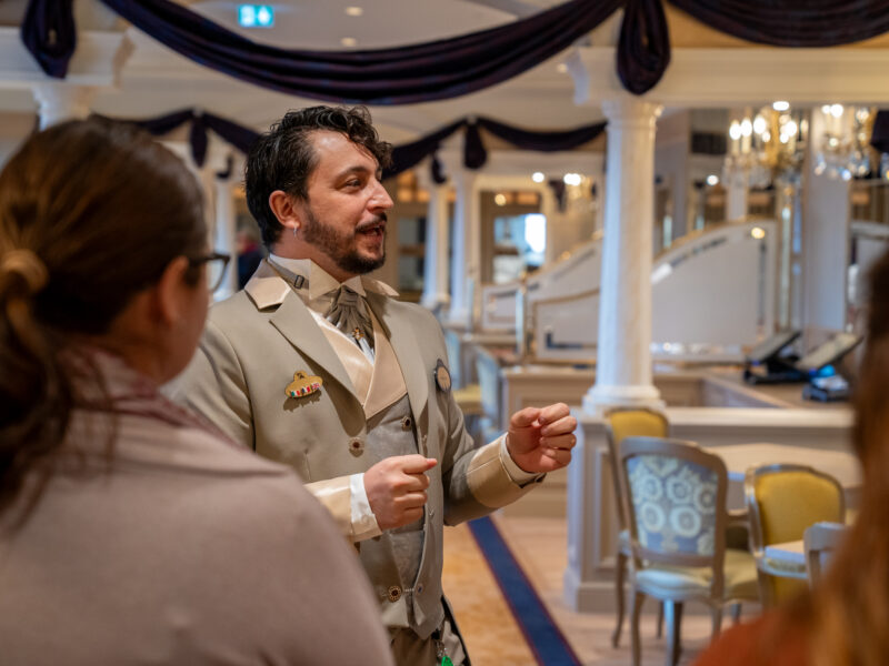 Un homme en costume beige s'adresse à deux personnes dans une salle décorée et élégante avec des colonnes, des chaises et des tissus drapés lors d'un événement de recrutement à Villeurbanne.