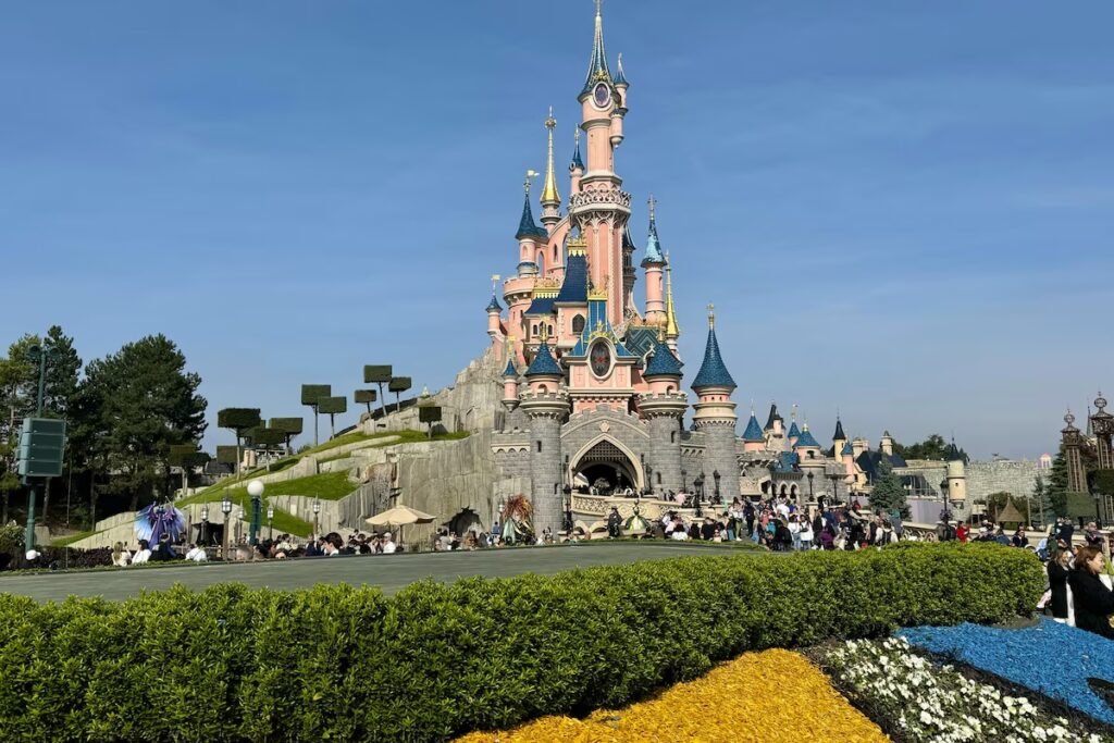 Un grand château rose aux flèches bleues se dresse au centre de l'un des célèbres parcs Disney, entouré de visiteurs et de parterres de fleurs colorés sous un ciel bleu limpide.