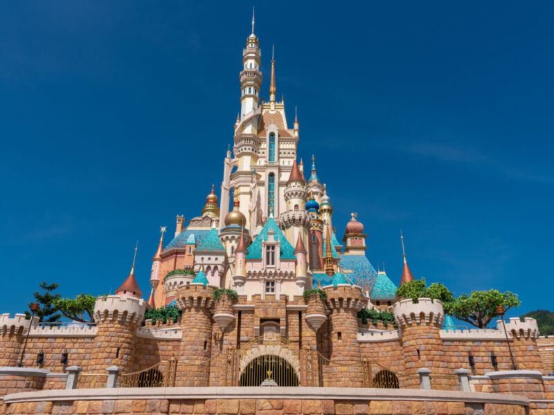 Un grand château coloré aux multiples flèches et tourelles sous un ciel bleu clair, entouré de murs de pierre et de verdure, rappelant l'atmosphère enchanteresse de Disneyland Thaïlande dans le Corridor Économique Oriental.