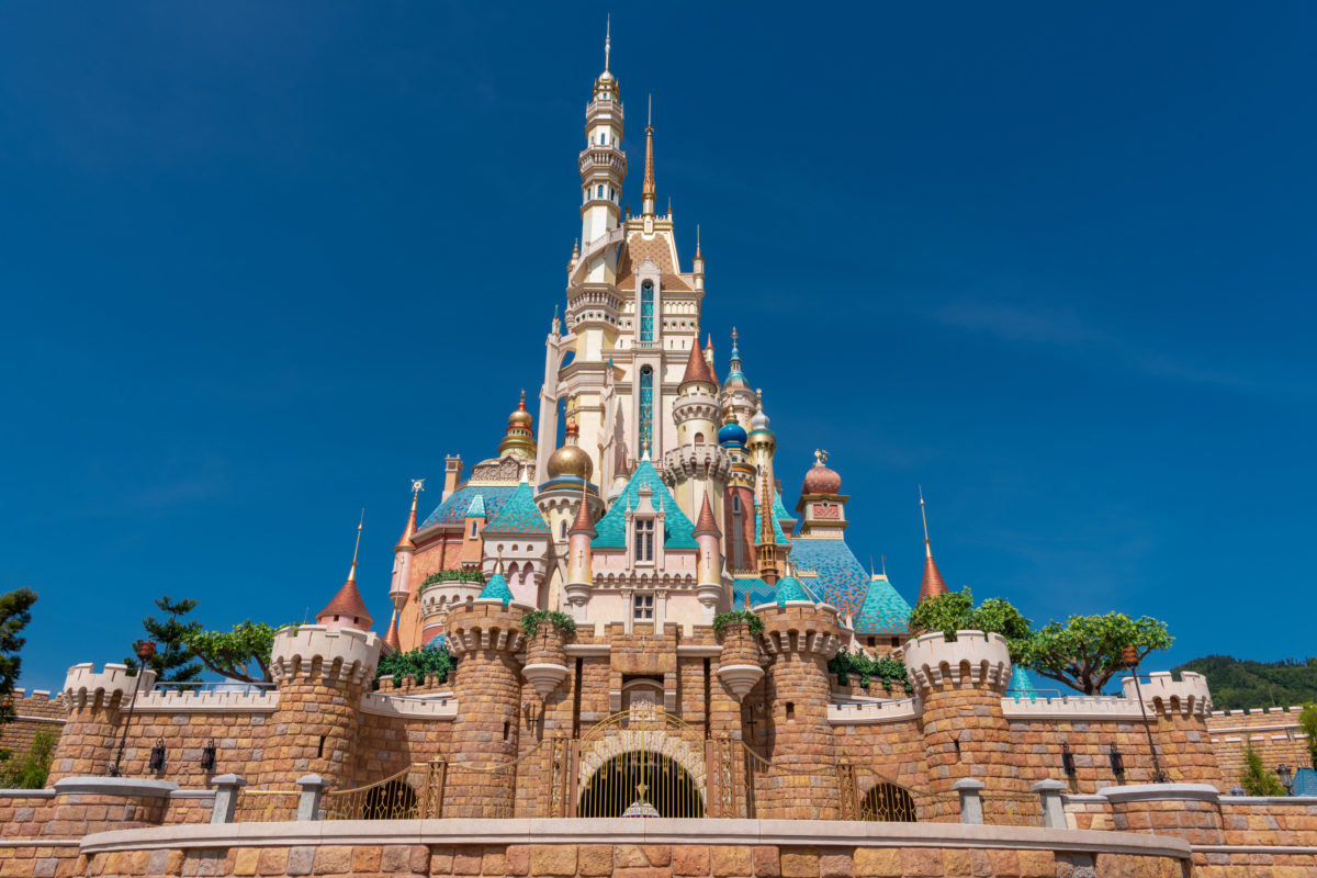 Un grand château coloré aux multiples flèches et tourelles sous un ciel bleu clair, entouré de murs de pierre et de verdure, rappelant l'atmosphère enchanteresse de Disneyland Thaïlande dans le Corridor Économique Oriental.
