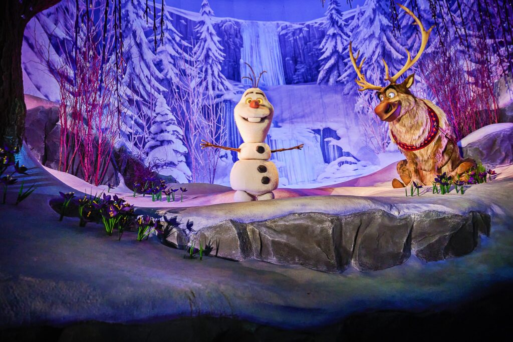 Olaf le bonhomme de neige et Sven le renne se trouvent sur une scène enneigée à Disney Adventure World, entourés de paysages hivernaux tels que des arbres couverts de neige et une cascade gelée en arrière-plan.