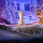 Olaf le bonhomme de neige et Sven le renne se trouvent sur une scène enneigée à Disney Adventure World, entourés de paysages hivernaux tels que des arbres couverts de neige et une cascade gelée en arrière-plan.