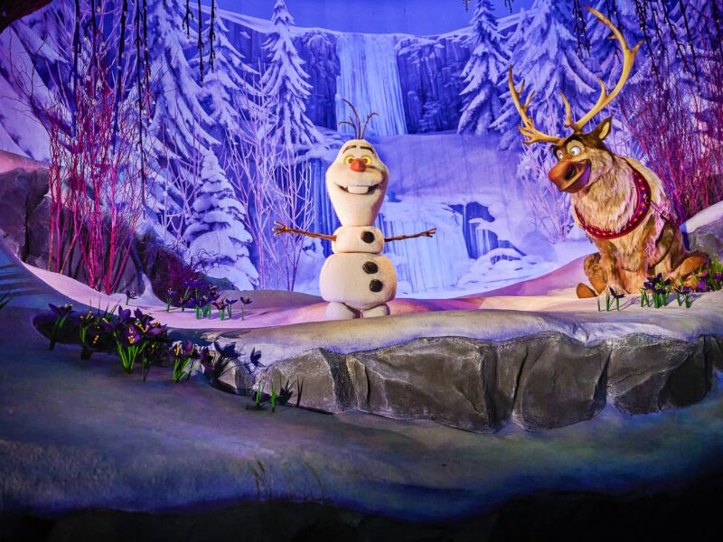 Olaf le bonhomme de neige et Sven le renne se trouvent sur une scène enneigée à Disney Adventure World, entourés de paysages hivernaux tels que des arbres couverts de neige et une cascade gelée en arrière-plan.