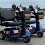 Deux scooters électriques de location bleus et orange portant l'inscription GMS sont garés côte à côte sur une allée pavée près d'un plan d'eau à Chalifert, non loin de Disneyland Paris.