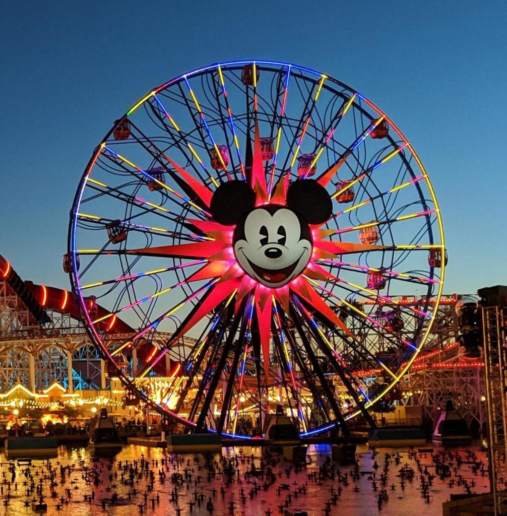 Une grande roue avec un visage de Mickey Mouse au centre est brillamment éclairée au crépuscule à Disney California Adventure, avec des personnes et des attractions du parc d'attractions visibles en dessous.