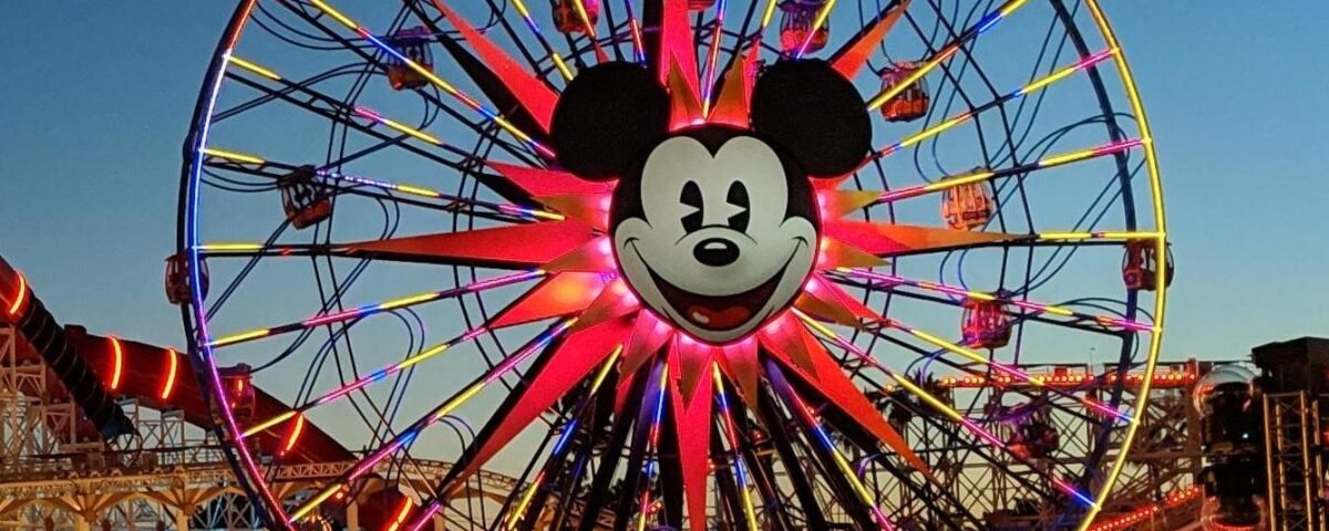 Une grande roue avec un visage de Mickey Mouse au centre est brillamment éclairée au crépuscule à Disney California Adventure, avec des personnes et des attractions du parc d'attractions visibles en dessous.
