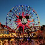 Une grande roue avec un visage de Mickey Mouse au centre est brillamment éclairée au crépuscule à Disney California Adventure, avec des personnes et des attractions du parc d'attractions visibles en dessous.