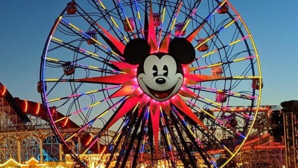 Une grande roue avec un visage de Mickey Mouse au centre est brillamment éclairée au crépuscule à Disney California Adventure, avec des personnes et des attractions du parc d'attractions visibles en dessous.