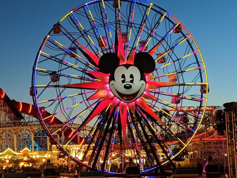 Une grande roue avec un visage de Mickey Mouse au centre est brillamment éclairée au crépuscule à Disney California Adventure, avec des personnes et des attractions du parc d'attractions visibles en dessous.