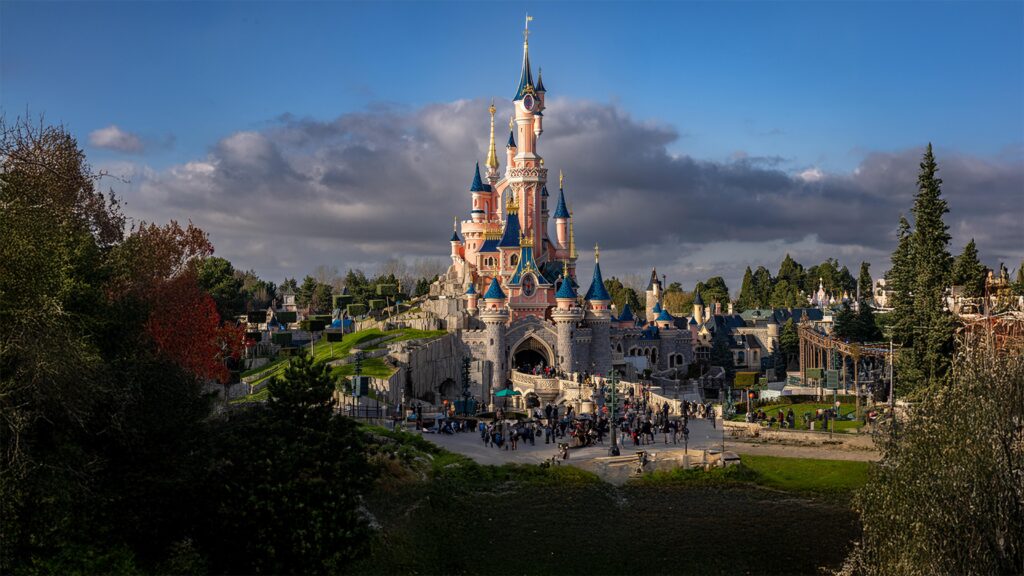 Un grand château de conte de fées aux flèches bleues se dresse au centre d'un parc à thème, mettant en valeur l'accessibilité de Disneyland Paris à tous les visiteurs, entouré de foule et de verdure sous un ciel partiellement nuageux.