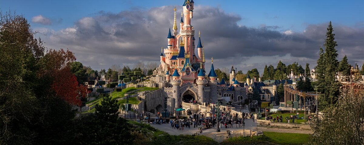 Un grand château de conte de fées aux flèches bleues se dresse au centre d'un parc à thème, mettant en valeur l'accessibilité de Disneyland Paris à tous les visiteurs, entouré de foule et de verdure sous un ciel partiellement nuageux.