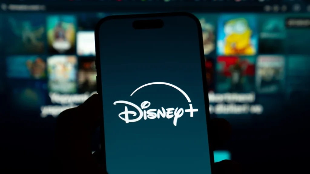 Un smartphone affiche le logo Disney+ en blanc sur un fond sombre, avec en arrière-plan un écran de télévision flou montrant divers contenus en streaming et des allusions aux innovations de la vidéo verticale du CES 2026.