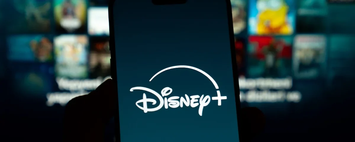 Un smartphone affiche le logo Disney+ en blanc sur un fond sombre, avec en arrière-plan un écran de télévision flou montrant divers contenus en streaming et des allusions aux innovations de la vidéo verticale du CES 2026.