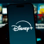 Un smartphone affiche le logo Disney+ en blanc sur un fond sombre, avec en arrière-plan un écran de télévision flou montrant divers contenus en streaming et des allusions aux innovations de la vidéo verticale du CES 2026.