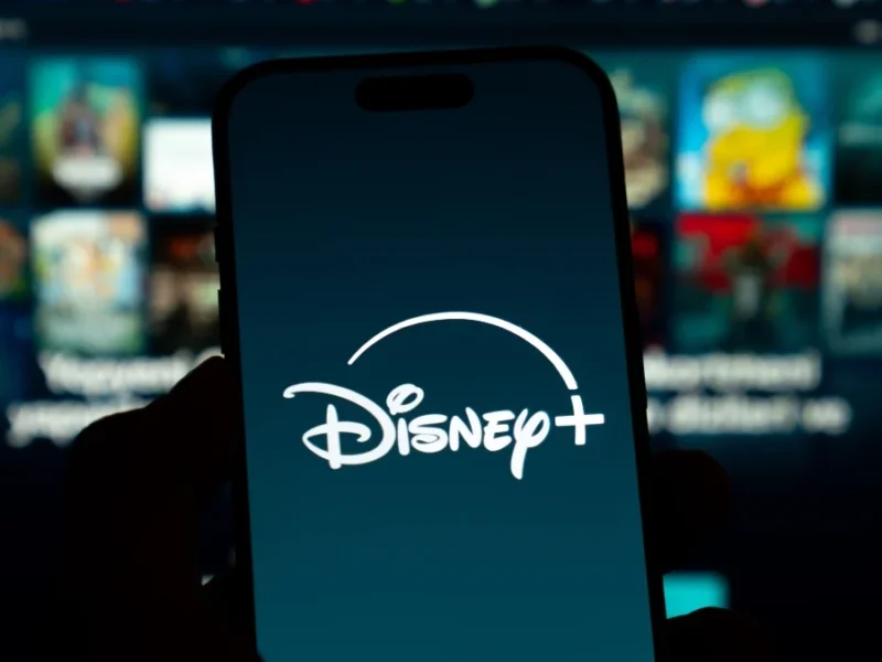 Un smartphone affiche le logo Disney+ en blanc sur un fond sombre, avec en arrière-plan un écran de télévision flou montrant divers contenus en streaming et des allusions aux innovations de la vidéo verticale du CES 2026.