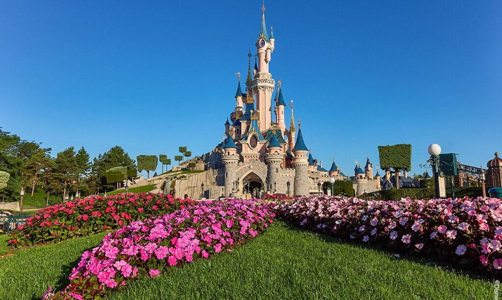 Un château aux tourelles bleues se dresse derrière des parterres de fleurs colorés et des pelouses vertes sous un ciel bleu limpide - une inspiration parfaite pour planifier une journée à Disneyland Paris.
