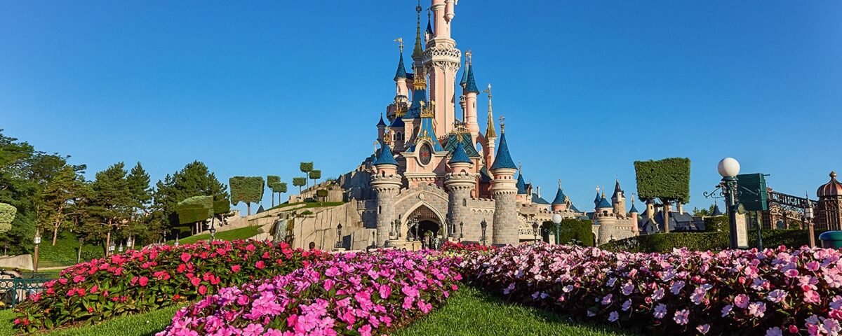 Un château aux tourelles bleues se dresse derrière des parterres de fleurs colorés et des pelouses vertes sous un ciel bleu limpide - une inspiration parfaite pour planifier une journée à Disneyland Paris.