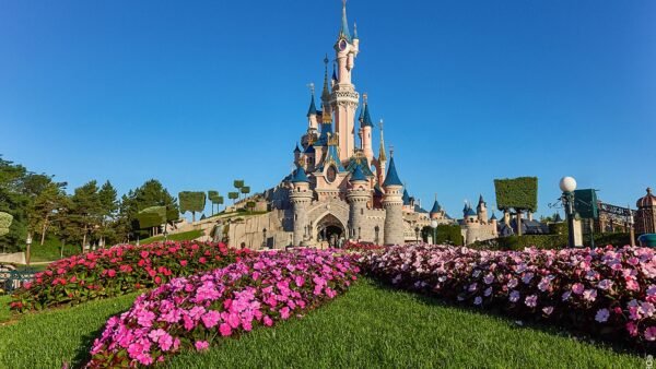 Un château aux tourelles bleues se dresse derrière des parterres de fleurs colorés et des pelouses vertes sous un ciel bleu limpide - une inspiration parfaite pour planifier une journée à Disneyland Paris.