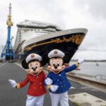 Deux personnes déguisées en Mickey et Minnie Mouse en uniforme de capitaine posent devant un grand bateau de croisière Disney Adventure amarré dans un port par une journée nuageuse.