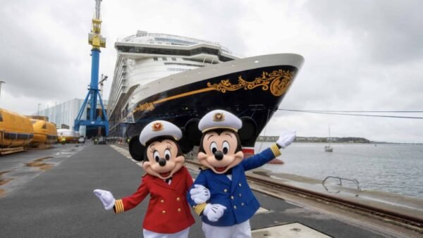 Deux personnes déguisées en Mickey et Minnie Mouse en uniforme de capitaine posent devant un grand bateau de croisière Disney Adventure amarré dans un port par une journée nuageuse.