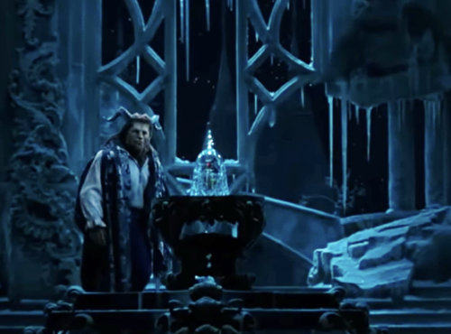 Un interprète costumé se tient à côté d'une rose en verre sur la scène, entouré de décors glacés de style gothique sous un éclairage bleu, évoquant la nostalgie des actions classiques en direct de Disney avec un approfondissement émotionnel.