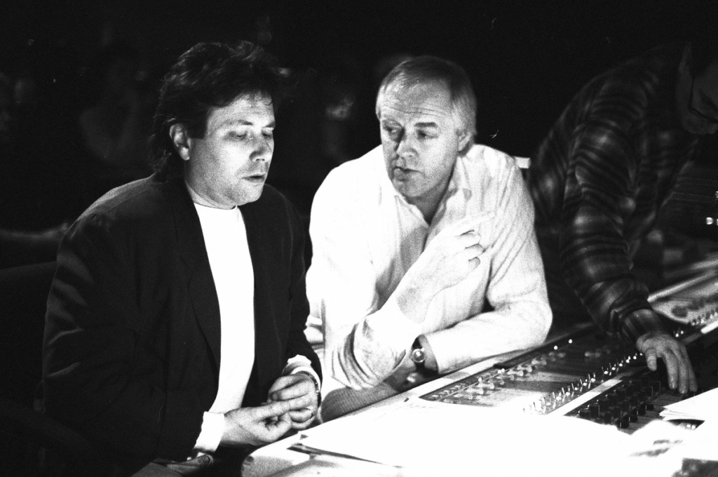 Deux hommes sont assis à une console de mixage audio, l'un parle et fait des gestes tandis que l'autre écoute attentivement, plongé dans un studio d'enregistrement empreint d'un sentiment de nostalgie.