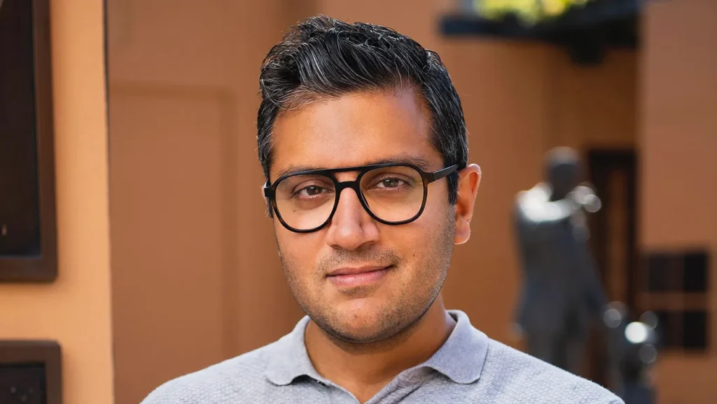 Un homme aux cheveux bruns courts et aux lunettes, ressemblant au directeur marketing de Disney, Asad Ayaz, se tient à l'extérieur, vêtu d'une chemise à col gris clair, sur un fond flou.