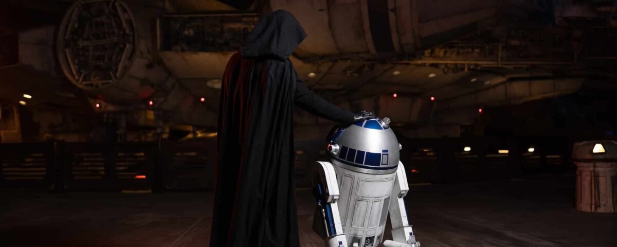 Une personne vêtue d'une cape noire, ressemblant à Dark Vador, se tient à côté du droïde R2-D2 devant un vaisseau spatial rappelant le Faucon Millenium à Galaxy's Edge.