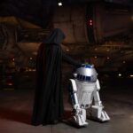 Une personne vêtue d'une cape noire, ressemblant à Dark Vador, se tient à côté du droïde R2-D2 devant un vaisseau spatial rappelant le Faucon Millenium à Galaxy's Edge.