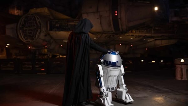 Une personne vêtue d'une cape noire, ressemblant à Dark Vador, se tient à côté du droïde R2-D2 devant un vaisseau spatial rappelant le Faucon Millenium à Galaxy's Edge.
